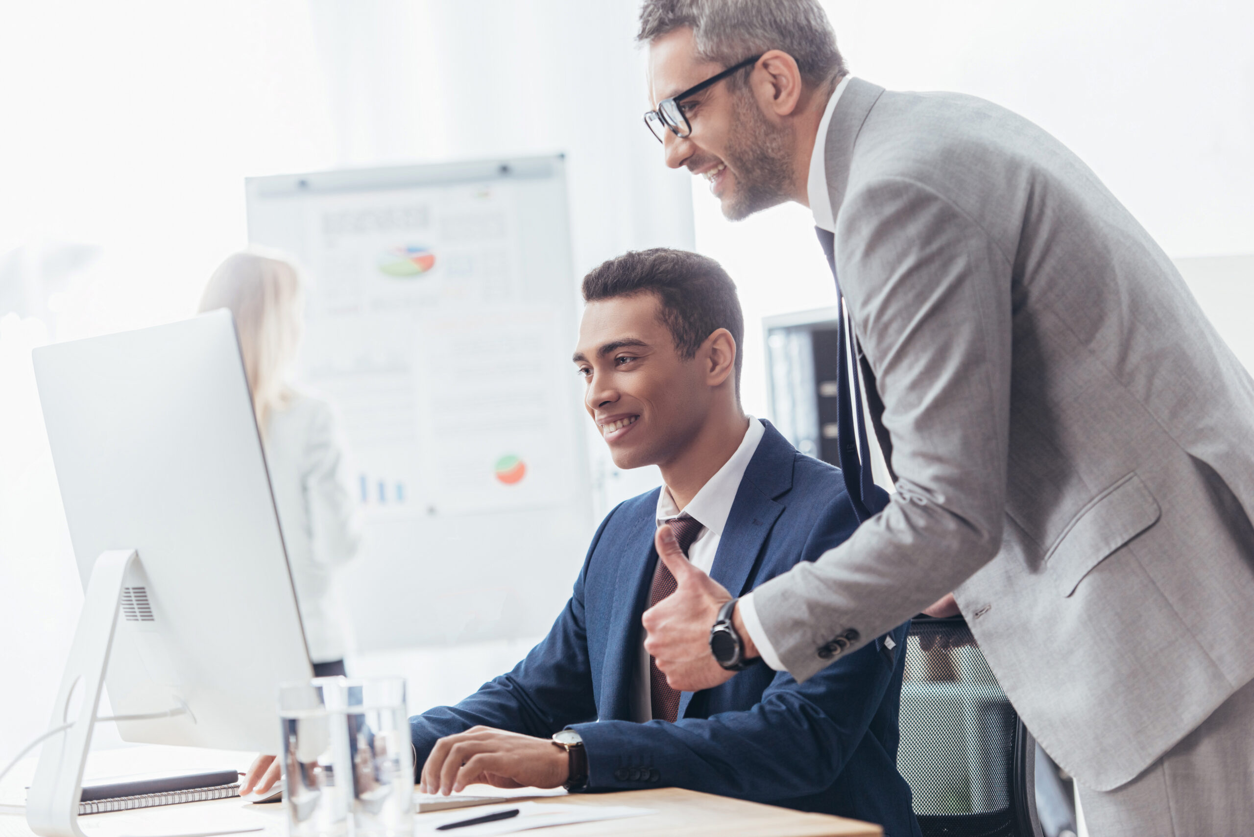 smiling businessman showing thumb up and looking at young colleague working with desktop computer