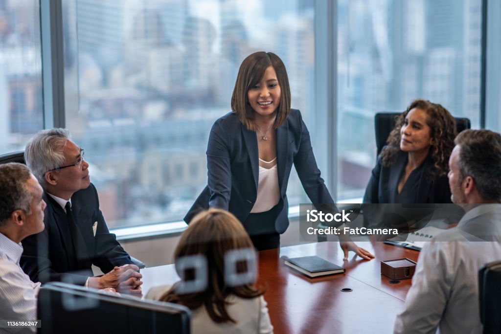 A group of adult businesspeople are indoors in an office. They are sitting around a table and having a meeting. One woman is leading the group.